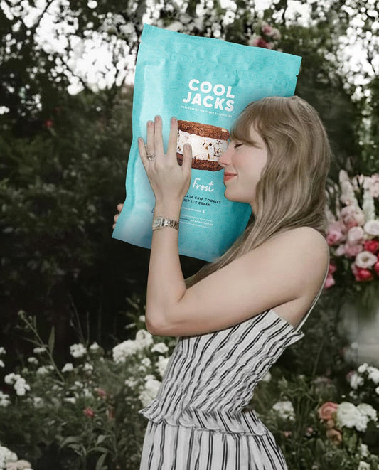 Taylor Swift smiling and holding a Cool Jacks Jack Frost ice cream sandwich pack in a flower garden.