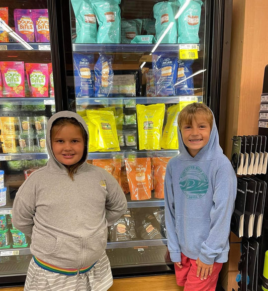 Two smiling kids in hoodies stand in front of a freezer filled with Cool Jacks ice cream sandwiches.